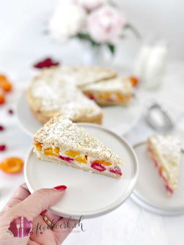 Himbeeren und Aprikosen Streuselkuchen auf weissem Teller serviert mit Hand im Bild